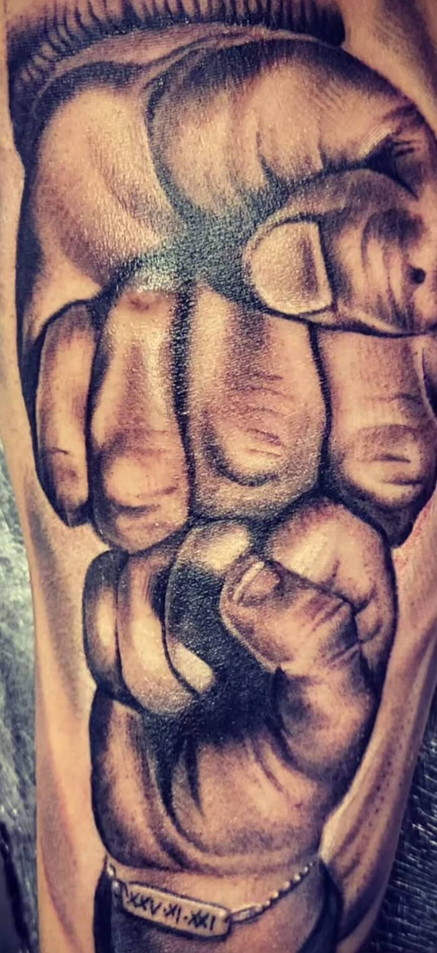 Black and grey realistic tattoo of two fists bumping — one adult hand and one small child’s hand — with detailed shading and a bracelet featuring Roman numerals, tattooed at Old Stone Tattoo House in Athlone, Ireland.
