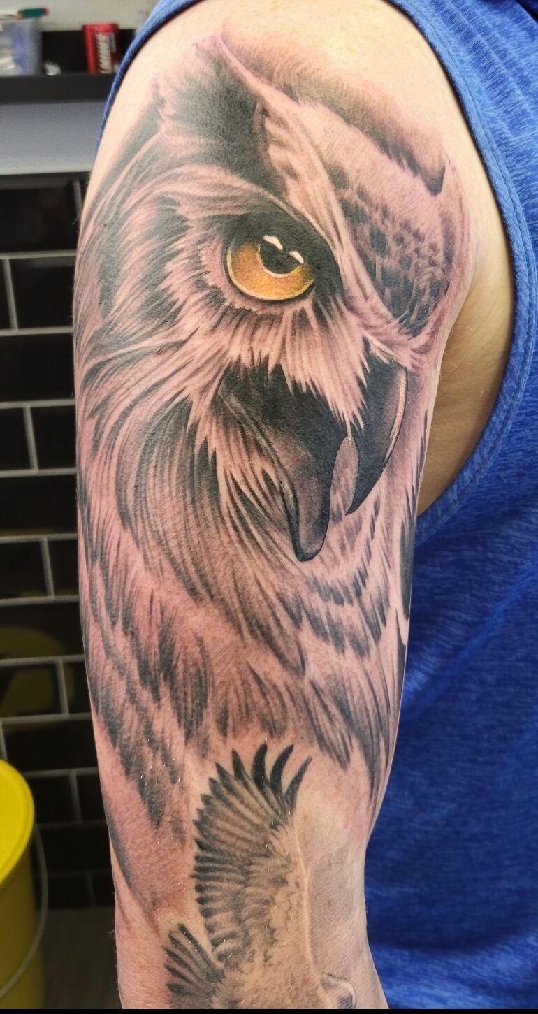 Realistic tattoo of a large owl’s face with piercing yellow-orange eye and sharp beak, complemented by a smaller flying owl below, inked in detailed black and grey shading with subtle color highlights