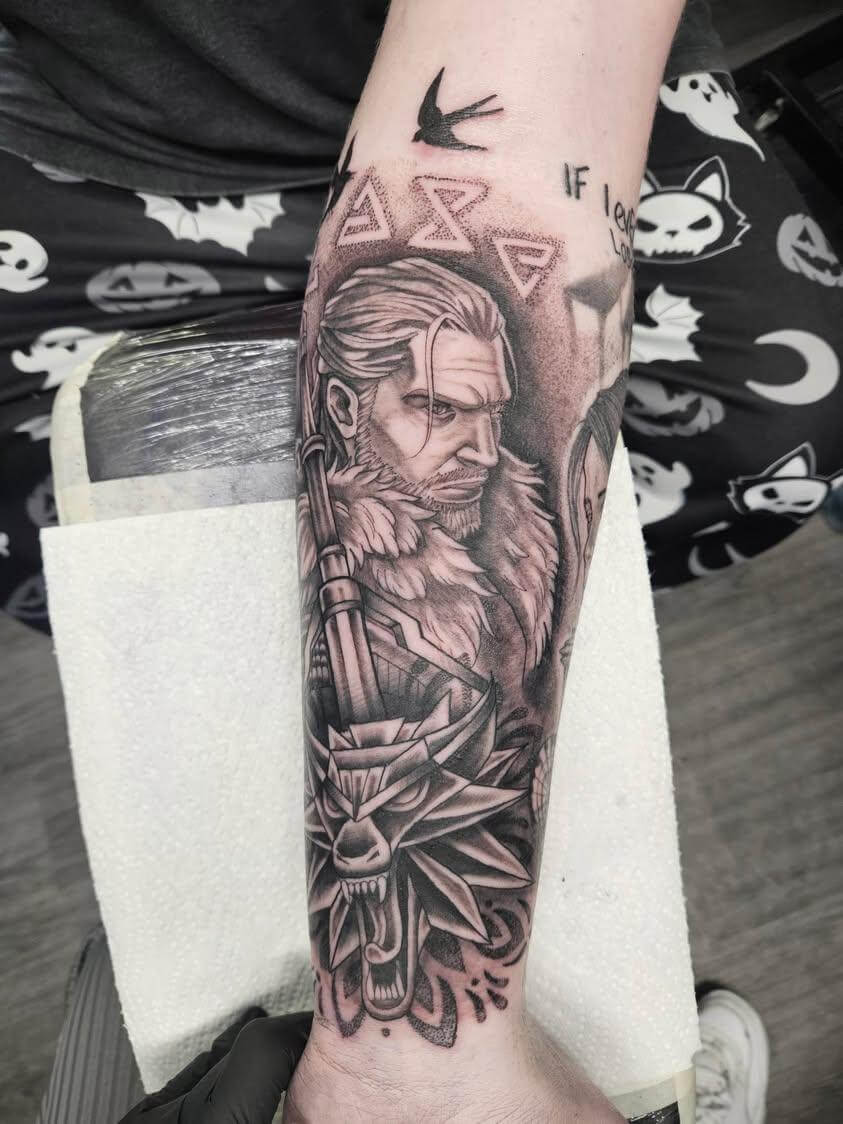 Black and grey tattoo of Geralt of Rivia from The Witcher, featuring his stern portrait with tied-back hair, fur collar, and a sword behind his head, above a detailed snarling wolf medallion design, with runic symbols and a small bird tattoo above.