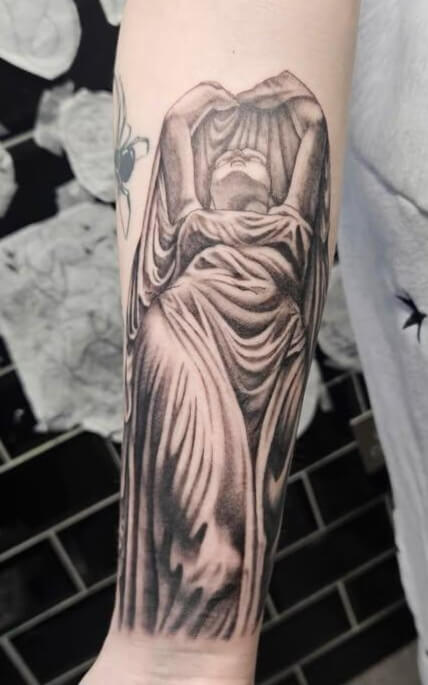 Black and grey tattoo of a robed, veiled figure with arms raised, draped in flowing cloth, resembling a classical statue carved in marble