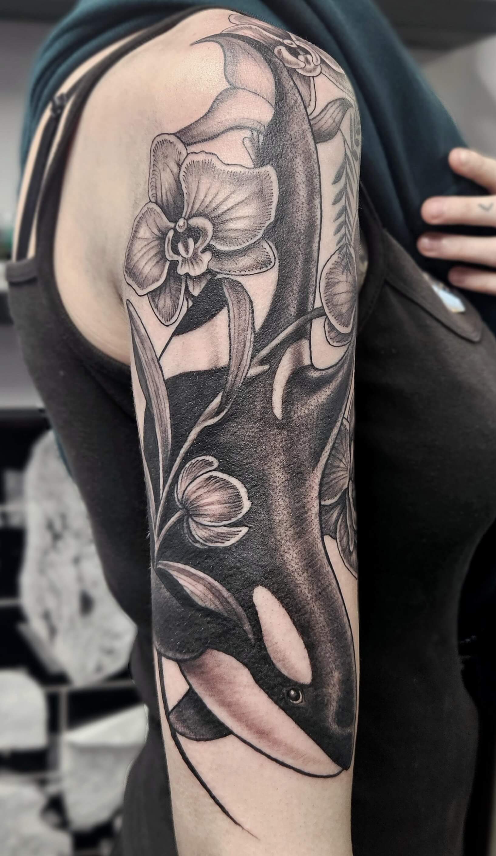 Black and grey tattoo of an orca swimming among detailed orchids and leaves, wrapping around the upper arm with intricate shading and smooth gradients.