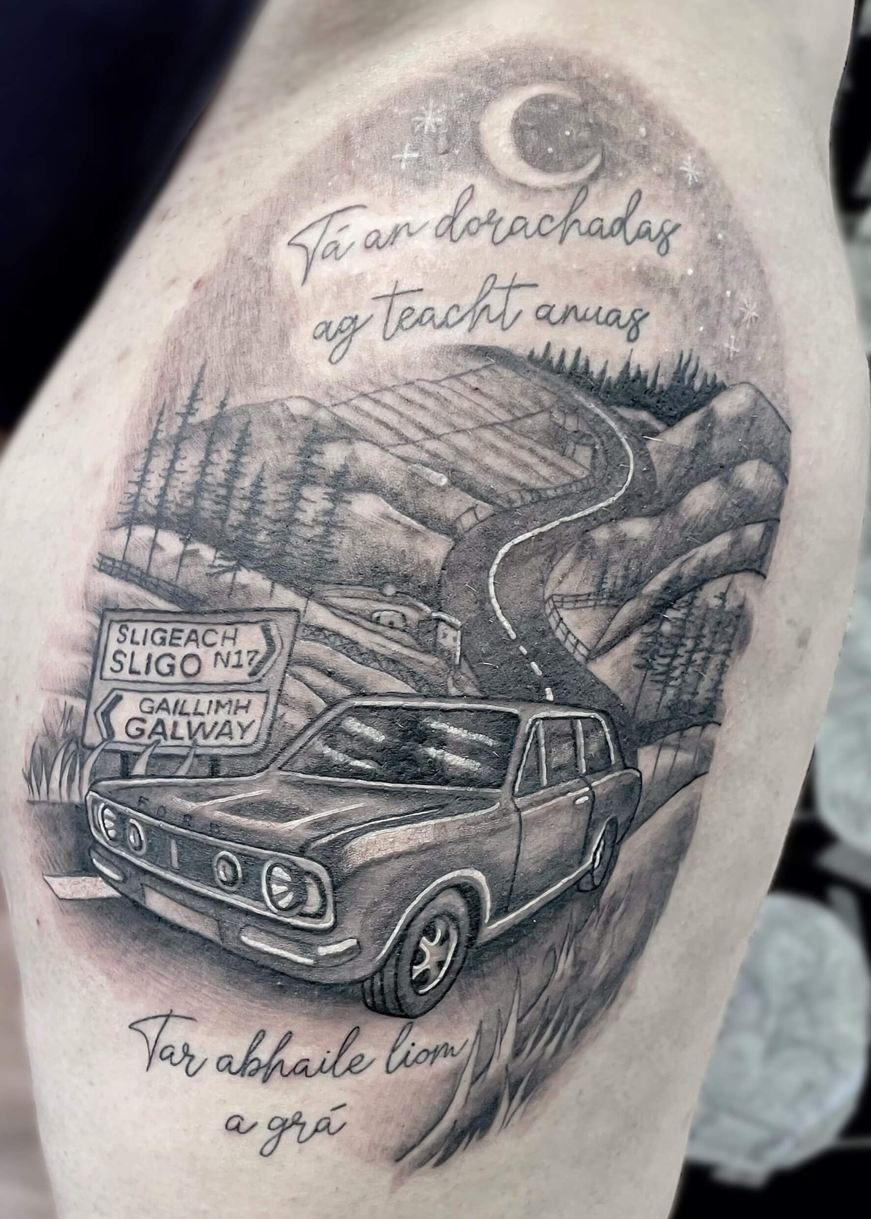 Black and grey tattoo of a vintage car on a winding country road under a crescent moon and starry night sky, with Irish road signs pointing to Sligo and Galway, and text in Irish above and below the scene