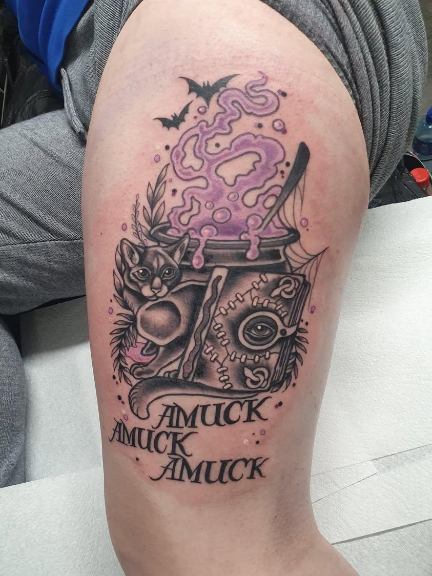 Black and grey thigh tattoo with purple highlights, showing a bubbling cauldron with rising smoke, a detailed spellbook with an eye, a cat, bats, and leaves, with bold lettering reading “Amuck Amuck Amuck.